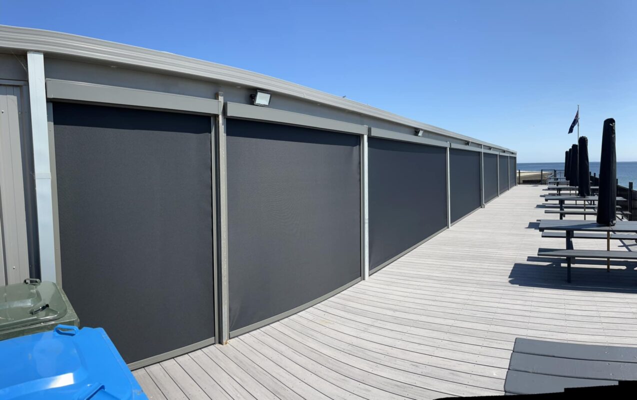 A run of charcoal Zipscreen outdoor blinds along a beachfront deck overlooking the bay
