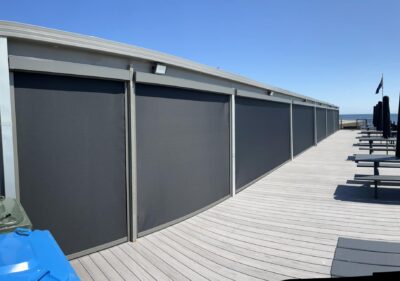 A run of charcoal Zipscreen outdoor blinds along a beachfront deck overlooking the bay